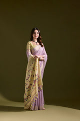 A woman models a Soft Muga Cotton Saree. The saree's body is light lavender, framed by a narrow Zari border. The pallu transitions into a pale yellow panel featuring colorful woven motifs. She pairs it with a light yellow blouse and a dark choker necklace.