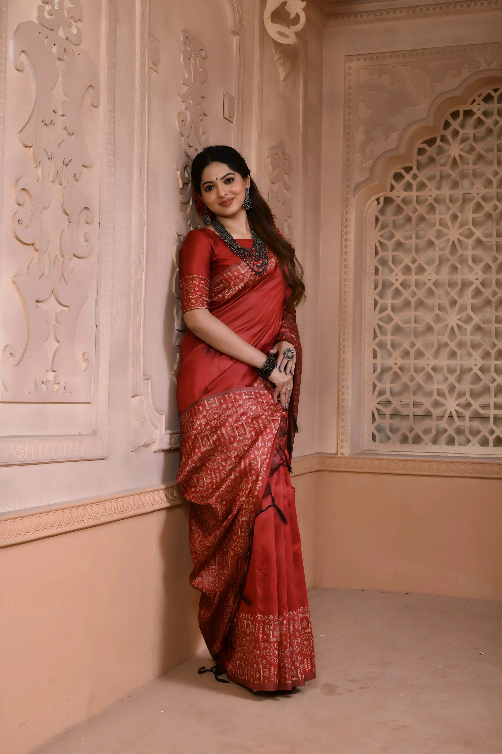  Woman wearing a maroon silk saree with intricate geometric border and pallu, paired with traditional jewelry, standing in a heritage-inspired room with ornate wall carvings and a decorative lattice window.