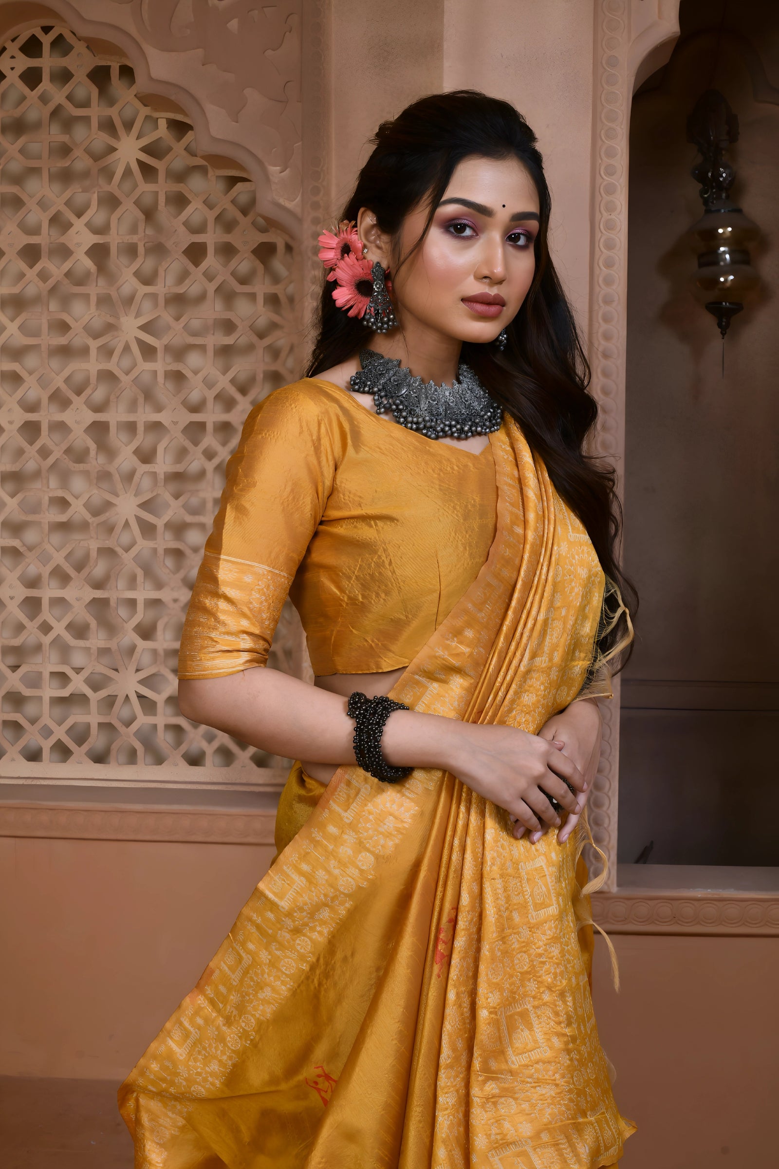 A woman models a vibrant Yellow Banglori Raw Silk Saree. The pallu and border feature dense, tone-on-tone Kalamkari-style weaving in gold Zari. She wears a yellow blouse and heavy dark gemstone jewelry, posing in an ornate, traditional setting.