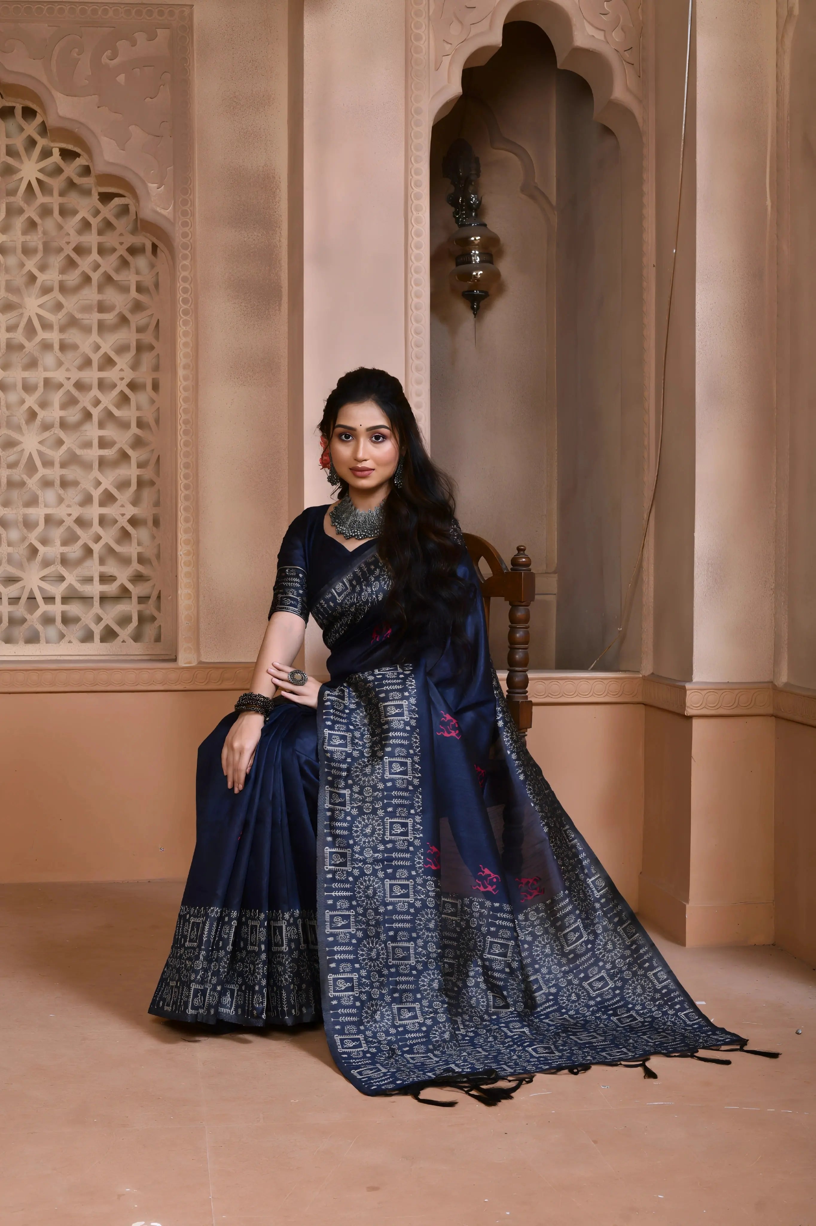 Woman wearing a navy blue silk saree with geometric patterned border and pallu, paired with traditional jewelry, standing in a heritage-inspired room with ornate carved windows and pastel walls