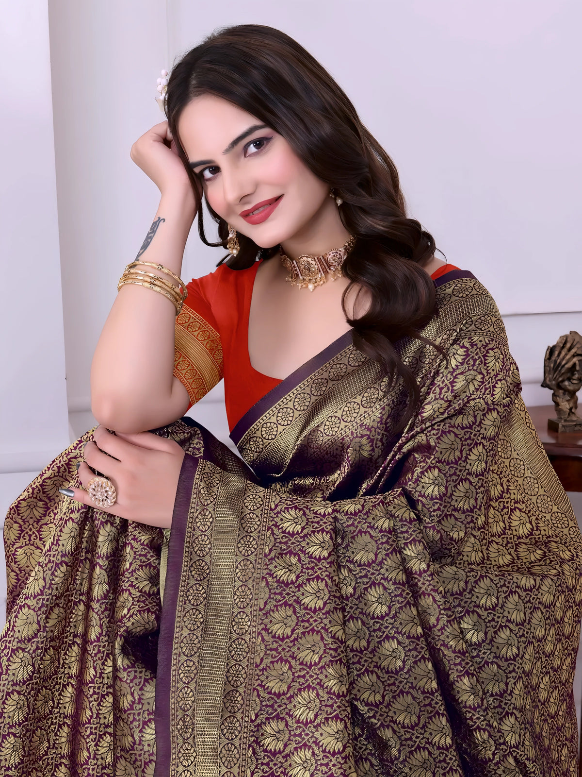 A woman models a striking Dark Brown Banarasi Silk Saree with dense all-over gold Zari weaving. The pallu provides a dramatic contrast in vibrant orange with heavy gold weaving. She wears an orange blouse and gold Kundan jewelry, posing indoors.