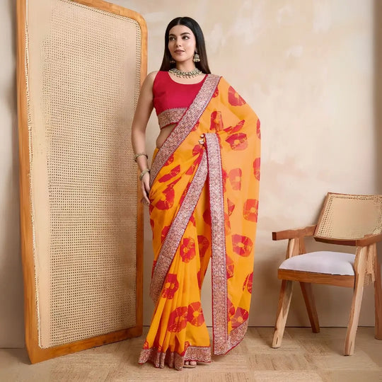 A woman models a vibrant Yellow Georgette Saree featuring an all-over bold red printed motif. The saree is bordered by dense metallic embroidery and beadwork. She wears a red sleeveless blouse and a heavy green gemstone or Kundan necklace.