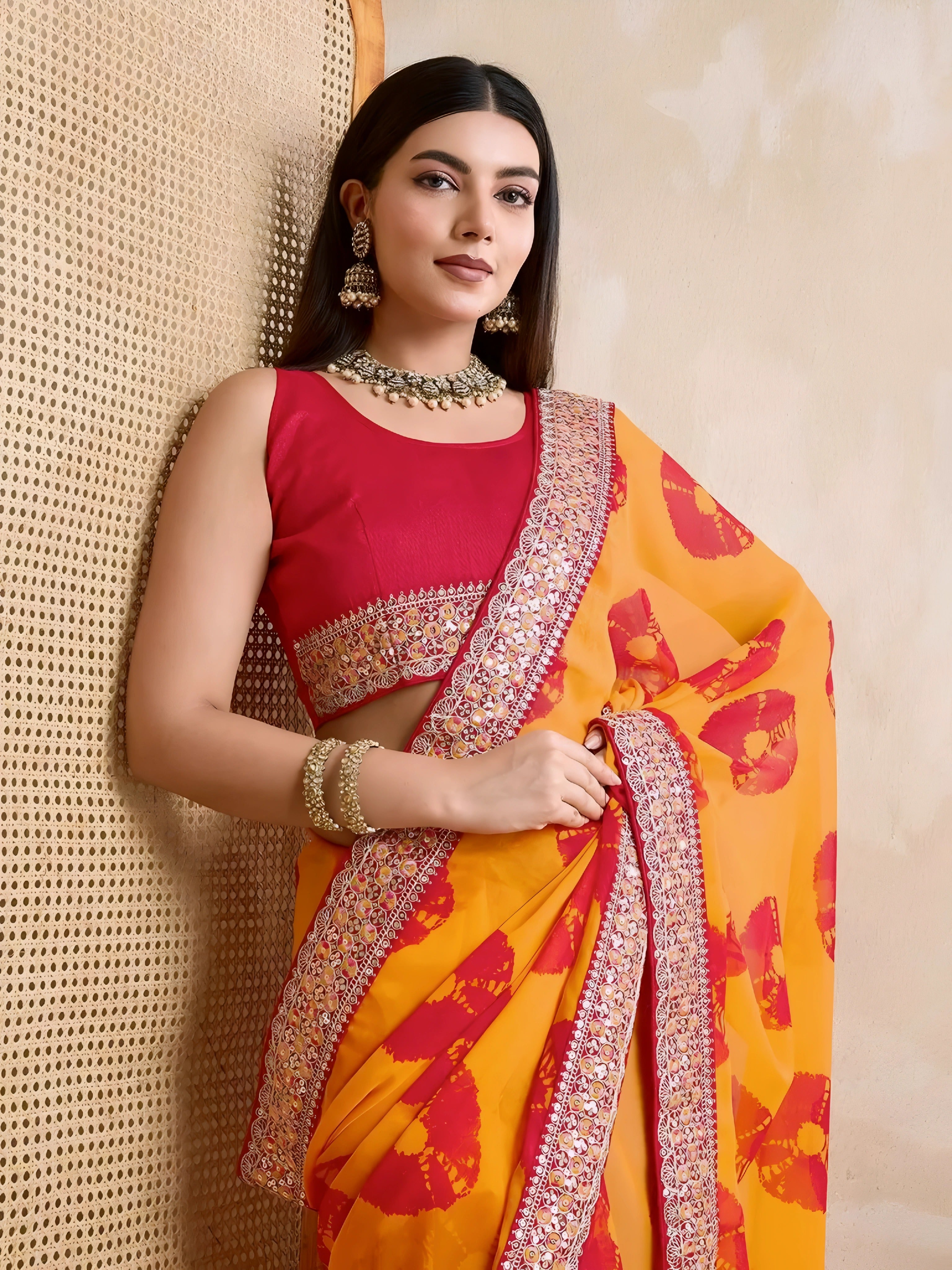 A woman models a vibrant Yellow Georgette Saree featuring an all-over bold red printed motif. The saree is bordered by dense metallic embroidery and beadwork. She wears a red sleeveless blouse and a heavy green gemstone or Kundan necklace.