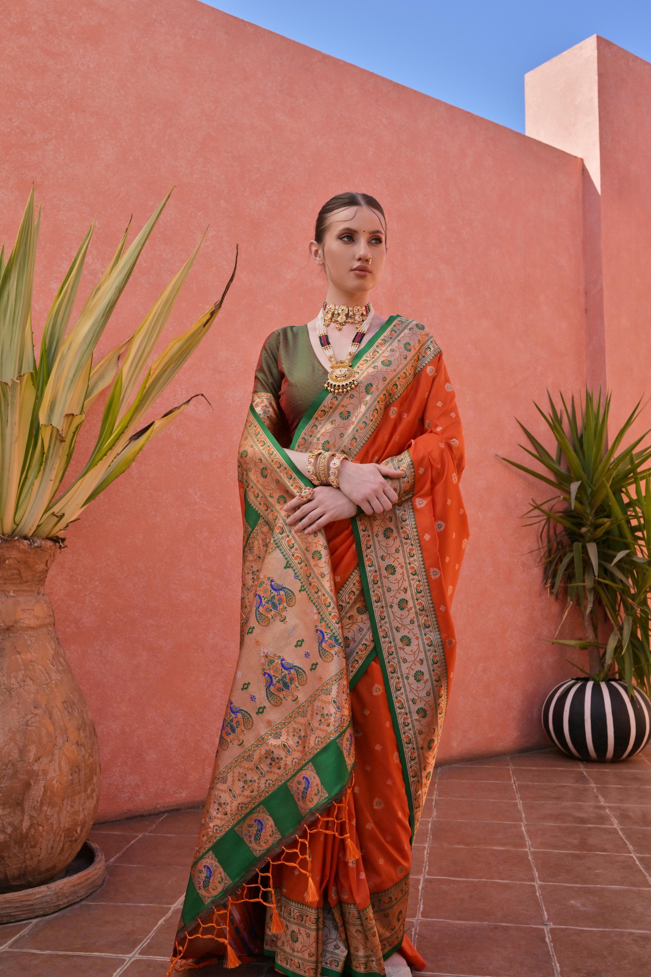 A woman models a vibrant Banarasi Silk Saree. The saree is deep orange with gold Zari weaving across the body, featuring a wide, ornate border and pallu with contrasting deep green accents. She pairs it with a short-sleeve green blouse and heavy traditional gold jewelry.