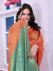 A woman models a striking Orange Banarasi Silk Saree with dense all-over gold Zari weaving. The pallu provides a spectacular contrast in sea green with heavy gold Banarasi weaving. She wears a sea green blouse and gold Kundan jewelry, posing by a window.