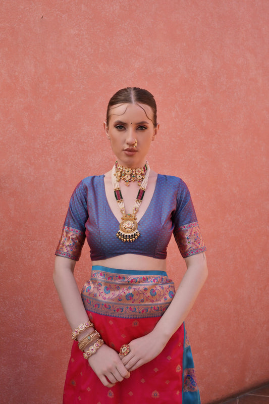A woman models a striking Soft Banarasi Silk Saree. The saree is deep pink with gold Zari weaving across the body, featuring an ornate border and pallu with contrasting blue accents. She pairs it with a short-sleeve blue blouse and heavy traditional gold jewelry.