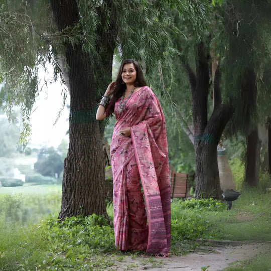 A woman models a Soft Tussar Silk Saree in a dusty rose pink color, covered in an all-over artistic print. The pallu features dense Zari weaving in a dark traditional pattern. She wears a pink blouse and a substantial set of oxidized silver jewelry, posing against a large tree.