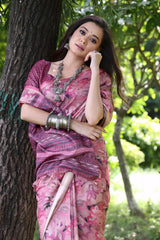 A woman models a Soft Tussar Silk Saree in a dusty rose pink color, covered in an all-over artistic print. The pallu features dense Zari weaving in a dark traditional pattern. She wears a pink blouse and a substantial set of oxidized silver jewelry, posing against a large tree.