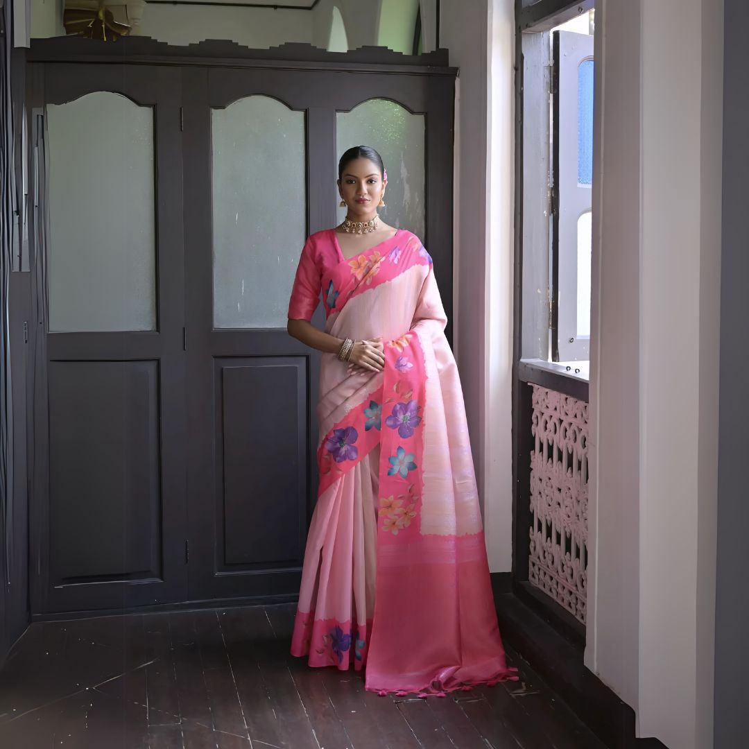 A woman models a graceful Soft Silk Saree. The saree body is pale pink with a wide hot pink border. The shoulder and pallu area feature large, vibrant printed floral motifs in blue and purple. She pairs it with a pink blouse and a gold choker necklace.