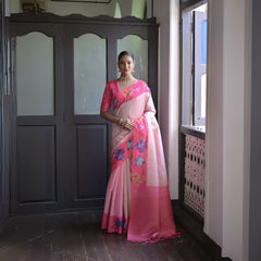 A woman models a graceful Soft Silk Saree. The saree body is pale pink with a wide hot pink border. The shoulder and pallu area feature large, vibrant printed floral motifs in blue and purple. She pairs it with a pink blouse and a gold choker necklace.