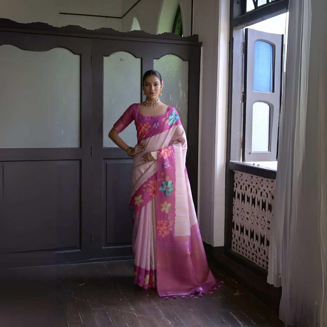 A woman models a graceful Soft Silk Saree. The saree body is light pink and features a wide, solid dark pink/fuchsia border. The shoulder and pallu area are decorated with large, vibrant printed floral motifs. She pairs it with a dark pink sheer-sleeve blouse and a gold choker necklace.