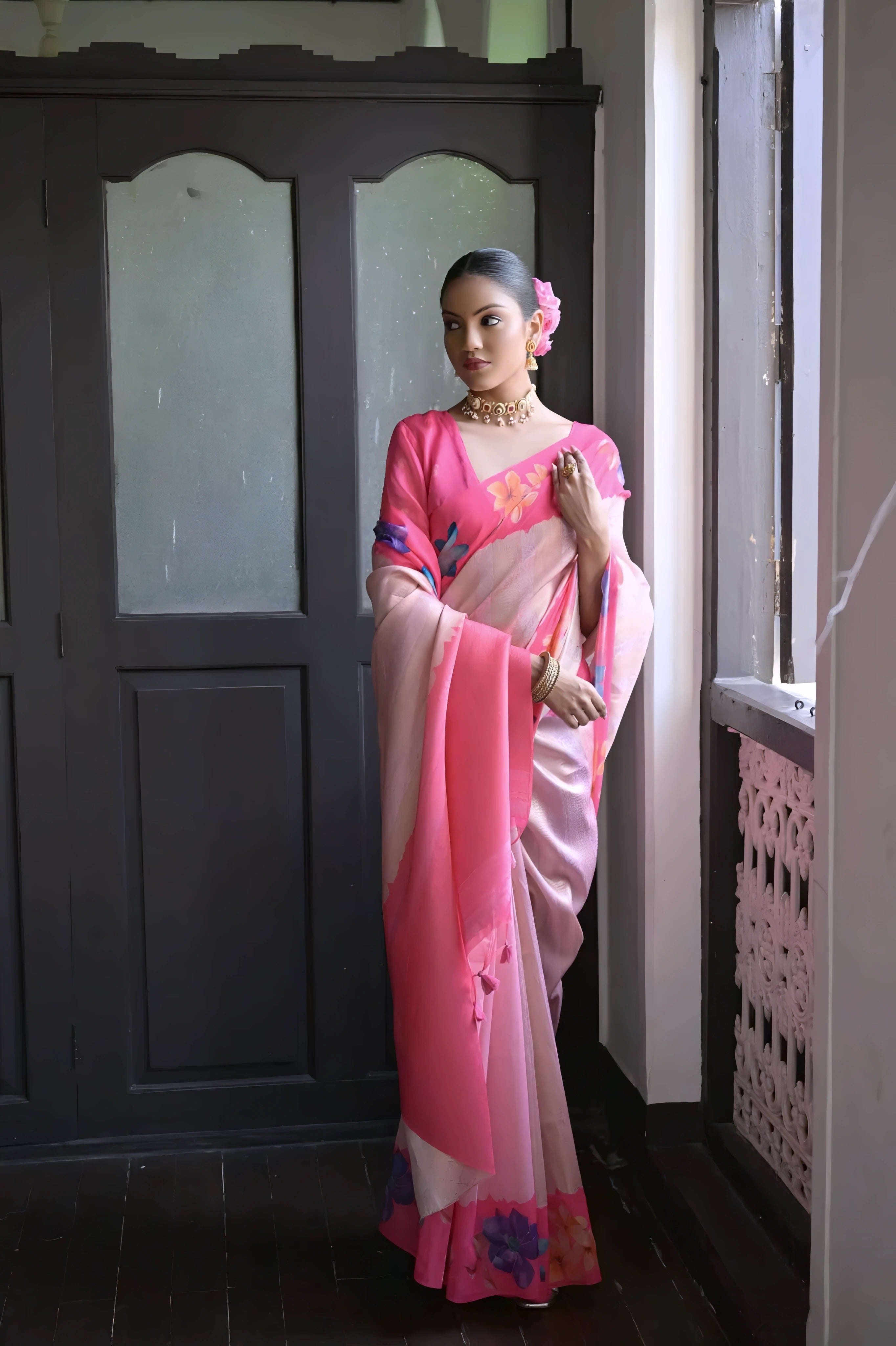 A woman models a graceful Soft Silk Saree. The saree body is pale pink with a wide hot pink border. The shoulder and pallu area feature large, vibrant printed floral motifs in blue and purple. She pairs it with a pink blouse and a gold choker necklace.