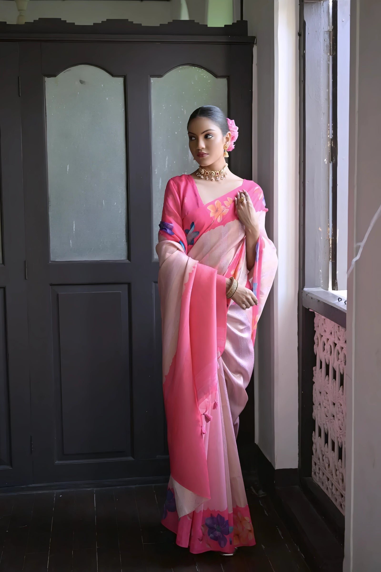 A woman models a graceful Soft Silk Saree. The saree body is pale pink with a wide hot pink border. The shoulder and pallu area feature large, vibrant printed floral motifs in blue and purple. She pairs it with a pink blouse and a gold choker necklace.