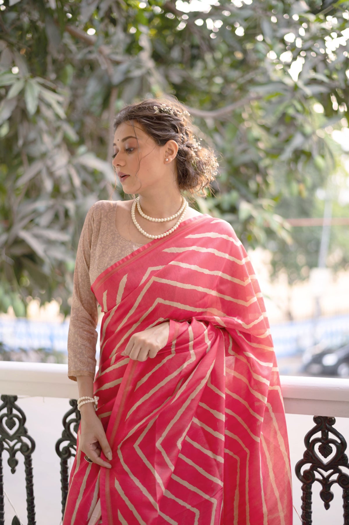 A woman models a vibrant Rose Pink Saree with a bold off-white chevron/striped pattern. The pallu is light beige with a dark border, featuring an elaborate printed motif of colorful flowers and foliage. She wears a pearl necklace and is posed in a doorway with green shutters.