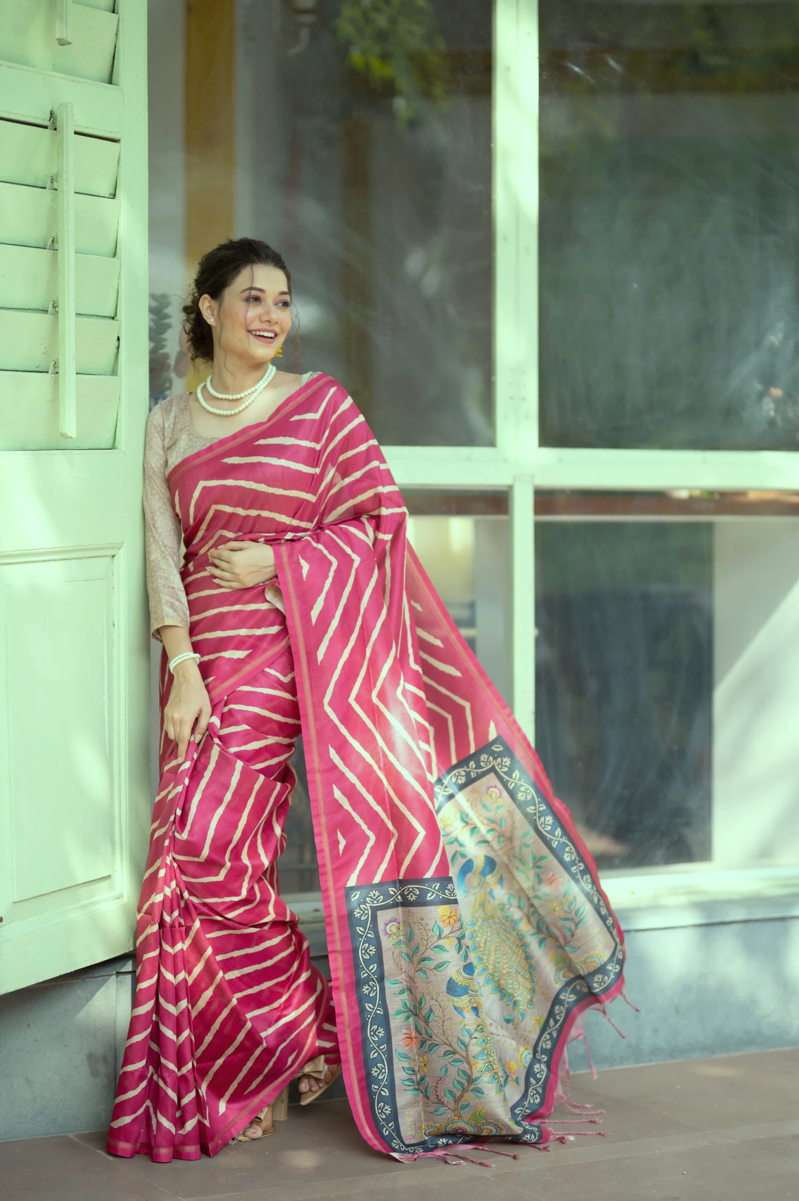 A woman models a vibrant Hot Pink Saree with a bold off-white chevron/striped pattern. The pallu is light beige with a dark border, featuring an elaborate printed motif of colorful flowers. She wears a pearl necklace and a large yellow flower hair accessory, posing indoors.