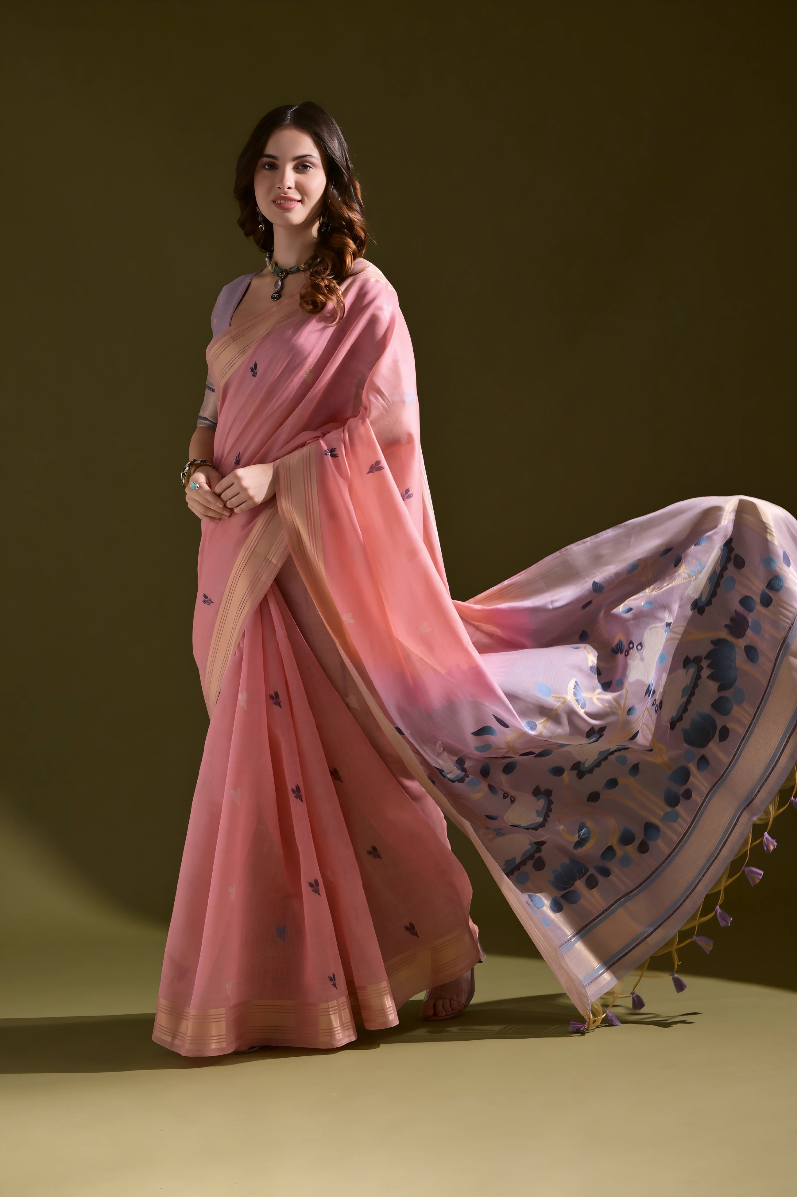 A woman models a Soft Muga Cotton Saree. The saree is soft pink with a gold Zari border. The pallu transitions into a pink and purple color scheme with large, woven blue and purple motifs. She pairs it with a light purple blouse and a choker necklace.