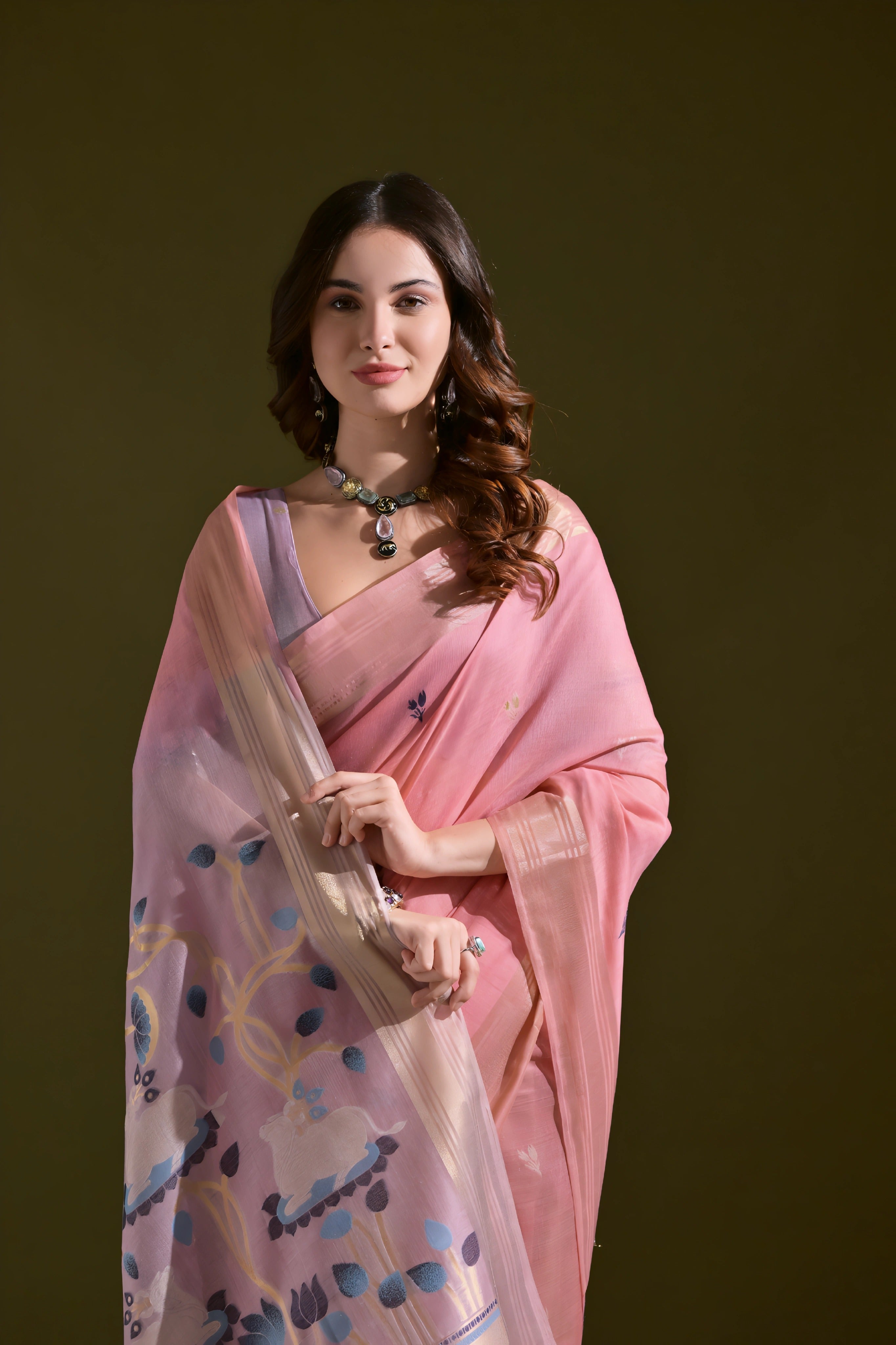 A woman models a Soft Muga Cotton Saree. The saree is soft pink with a gold Zari border. The pallu transitions into a pink and purple color scheme with large, woven blue and purple motifs. She pairs it with a light purple blouse and a choker necklace.