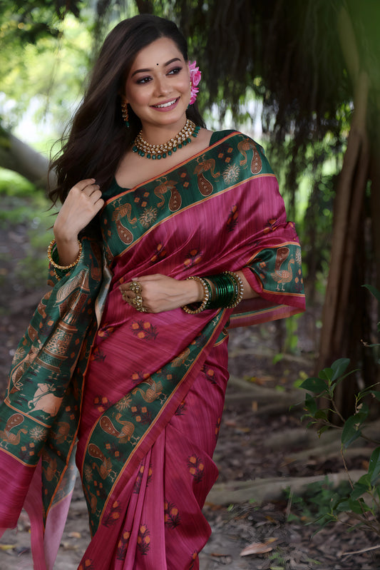 A woman models a Dark Pink Soft Tussar Silk Saree with small gold motifs. The saree has a wide, contrasting deep forest green border and pallu featuring intricate traditional and figurative prints. She wears a green blouse and is adorned with gold jewelry and a pink flower in her hair.