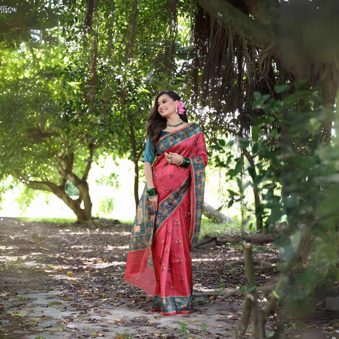 A woman models a vibrant Soft Tussar Silk Saree. The saree is maroon/deep rose pink with a wide, contrasting deep teal border and pallu featuring intricate traditional prints. She wears a teal blouse, a green beaded necklace, and is posed in a garden setting.