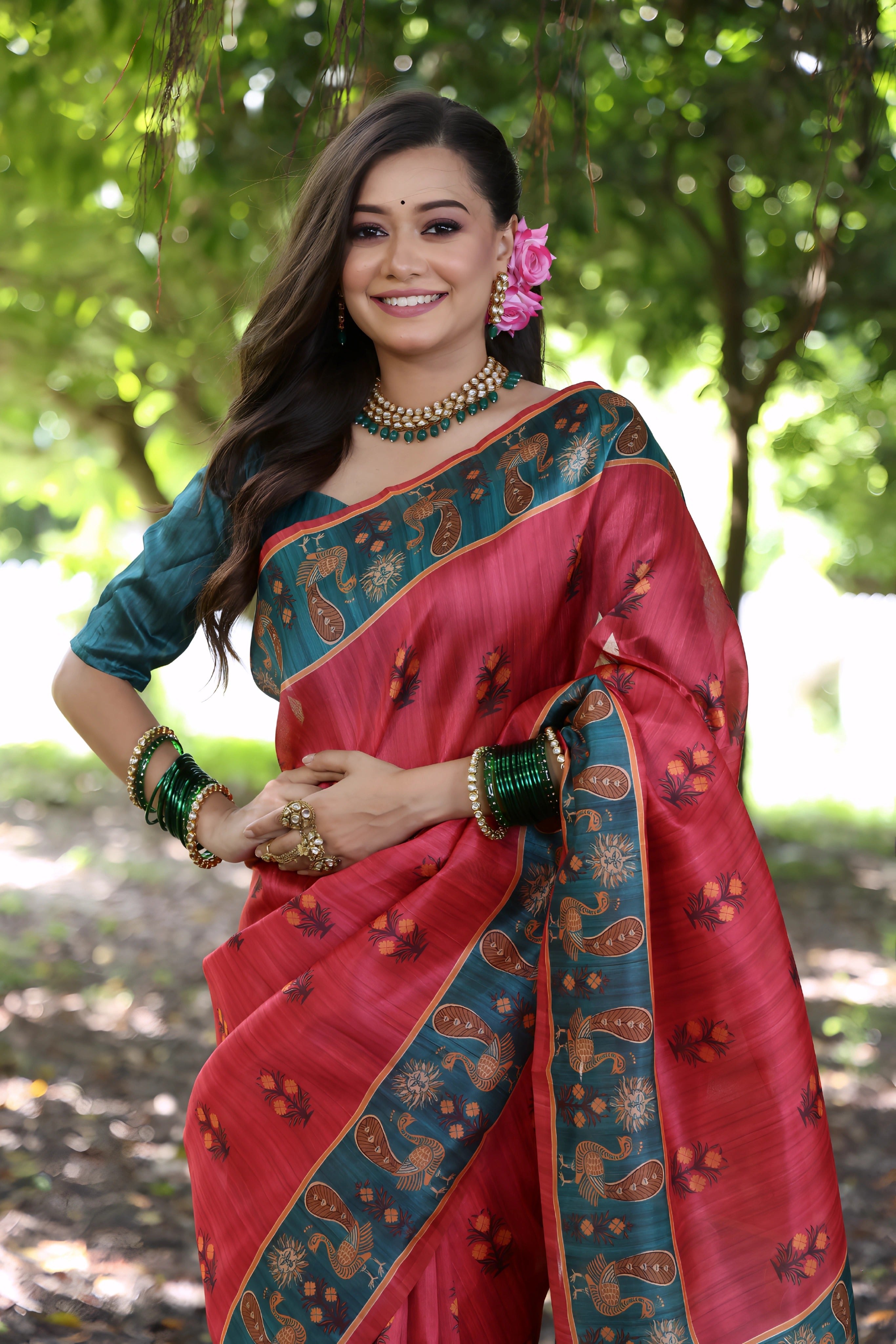 A woman models a vibrant Soft Tussar Silk Saree. The saree is maroon/deep rose pink with a wide, contrasting deep teal border and pallu featuring intricate traditional prints. She wears a teal blouse, a green beaded necklace, and is posed in a garden setting.