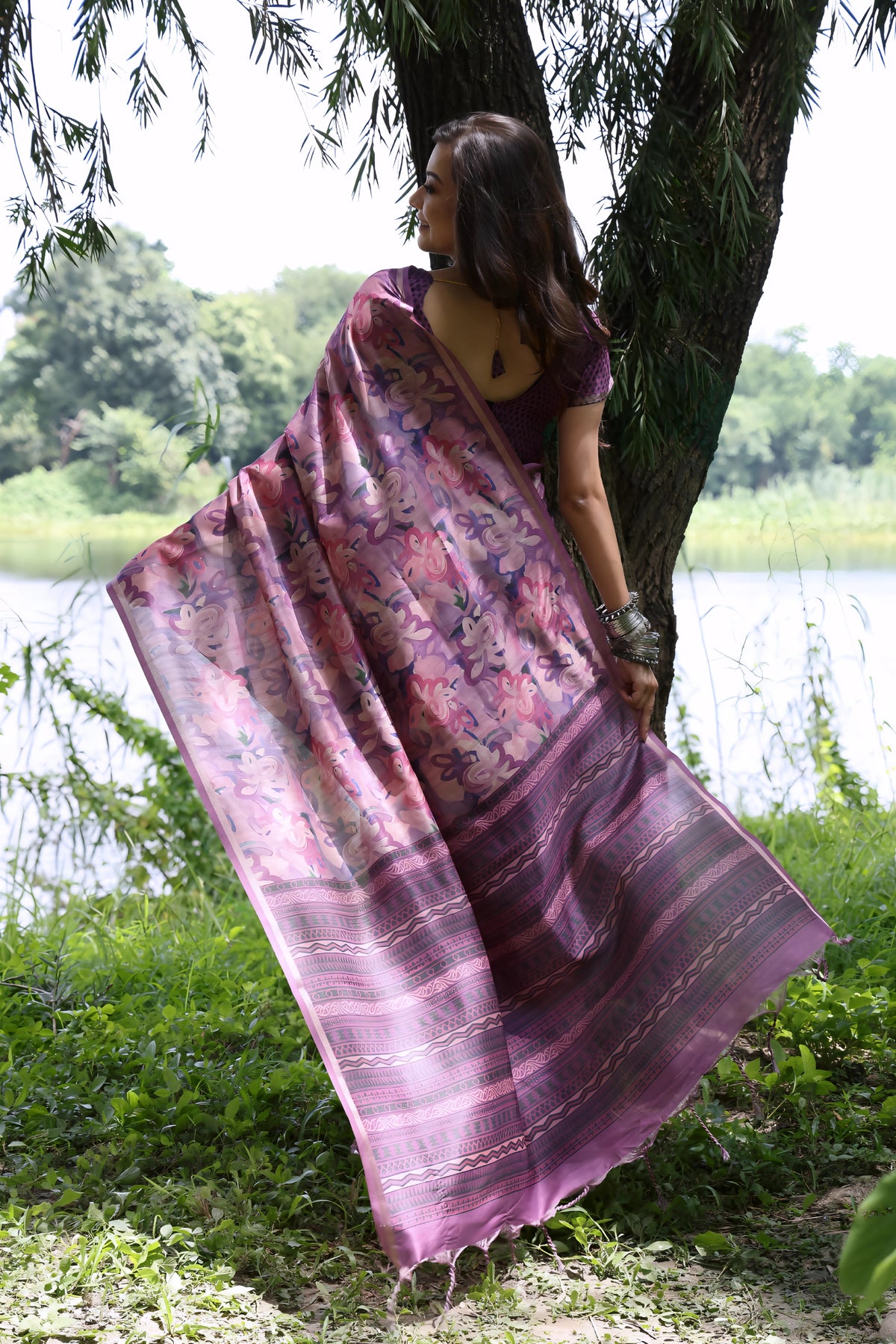 A woman models a Soft Tussar Silk Saree in dusty purple, covered in an elaborate all-over artistic print. The pallu features dense Zari weaving with a striped pattern. She wears a purple blouse and a substantial set of oxidized silver jewelry, posing against a large tree.