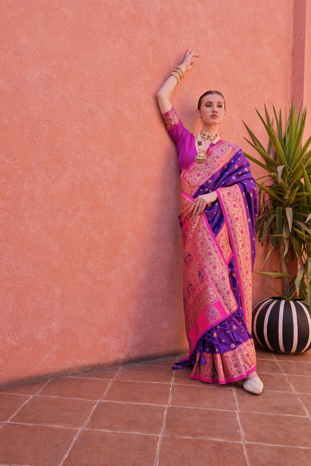 A woman models a vibrant Banarasi Silk Saree. The saree is royal purple with gold Zari weaving across the body, featuring a wide, ornate hot pink and gold Zari woven border and pallu. She pairs it with a short-sleeve pink blouse and heavy traditional gold jewelry.