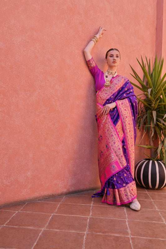 A woman models a vibrant Banarasi Silk Saree. The saree is royal purple with gold Zari weaving across the body, featuring a wide, ornate hot pink and gold Zari woven border and pallu. She pairs it with a short-sleeve pink blouse and heavy traditional gold jewelry.