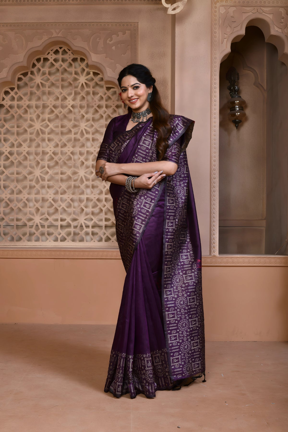 A woman models a striking Royal Purple Banglori Raw Silk Saree. The pallu and border feature dense, traditional Kalamkari-style weaving in metallic Zari thread. She wears a purple blouse and heavy green beaded jewelry, posing in an ornate, traditional setting.