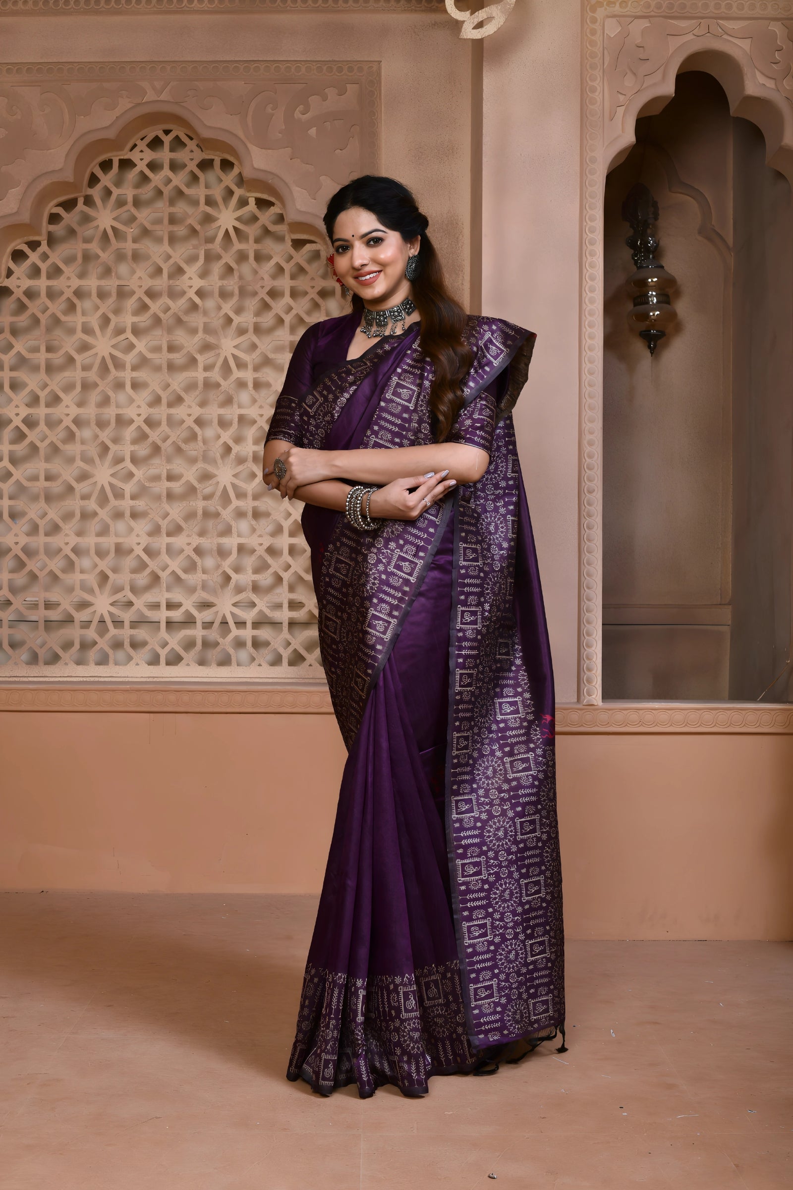 A woman models a striking Royal Purple Banglori Raw Silk Saree. The pallu and border feature dense, traditional Kalamkari-style weaving in metallic Zari thread. She wears a purple blouse and heavy green beaded jewelry, posing in an ornate, traditional setting.