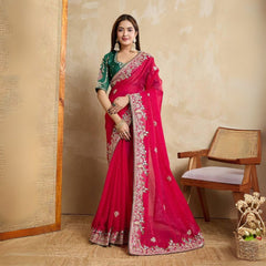 A woman models a vibrant Dark Pink Chiffon Silk Saree with scattered embellishments. The saree has a wide, intricate silver embroidery border. She wears a heavily embroidered dark green blouse and accessorizes with silver jewelry, posing indoors.