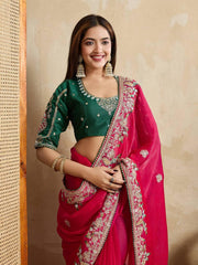 A woman models a vibrant Dark Pink Chiffon Silk Saree with scattered embellishments. The saree has a wide, intricate silver embroidery border. She wears a heavily embroidered dark green blouse and accessorizes with silver jewelry, posing indoors.