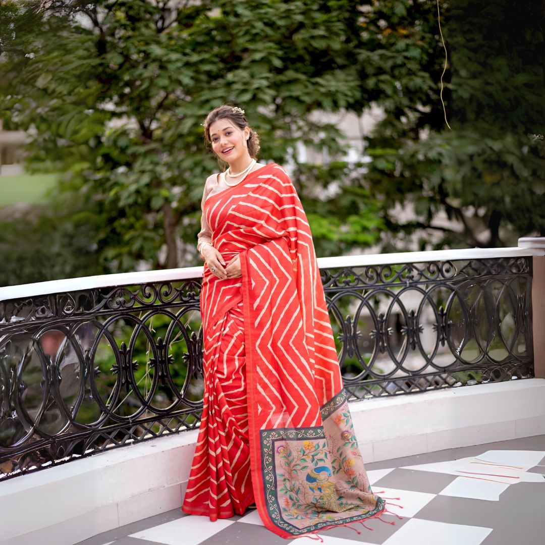 A woman models a vibrant Red Saree with a chevron/striped pattern. The pallu is beige with a dark green border, featuring elaborate printed motifs of birds and flowers. She wears a pearl necklace and is posed on an outdoor balcony.