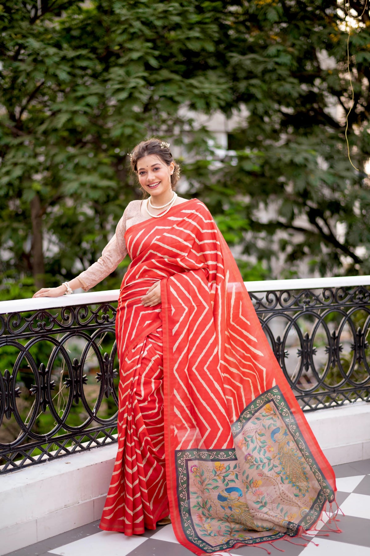 A woman models a vibrant Red Saree with a chevron/striped pattern. The pallu is beige with a dark green border, featuring elaborate printed motifs of birds and flowers. She wears a pearl necklace and is posed on an outdoor balcony.