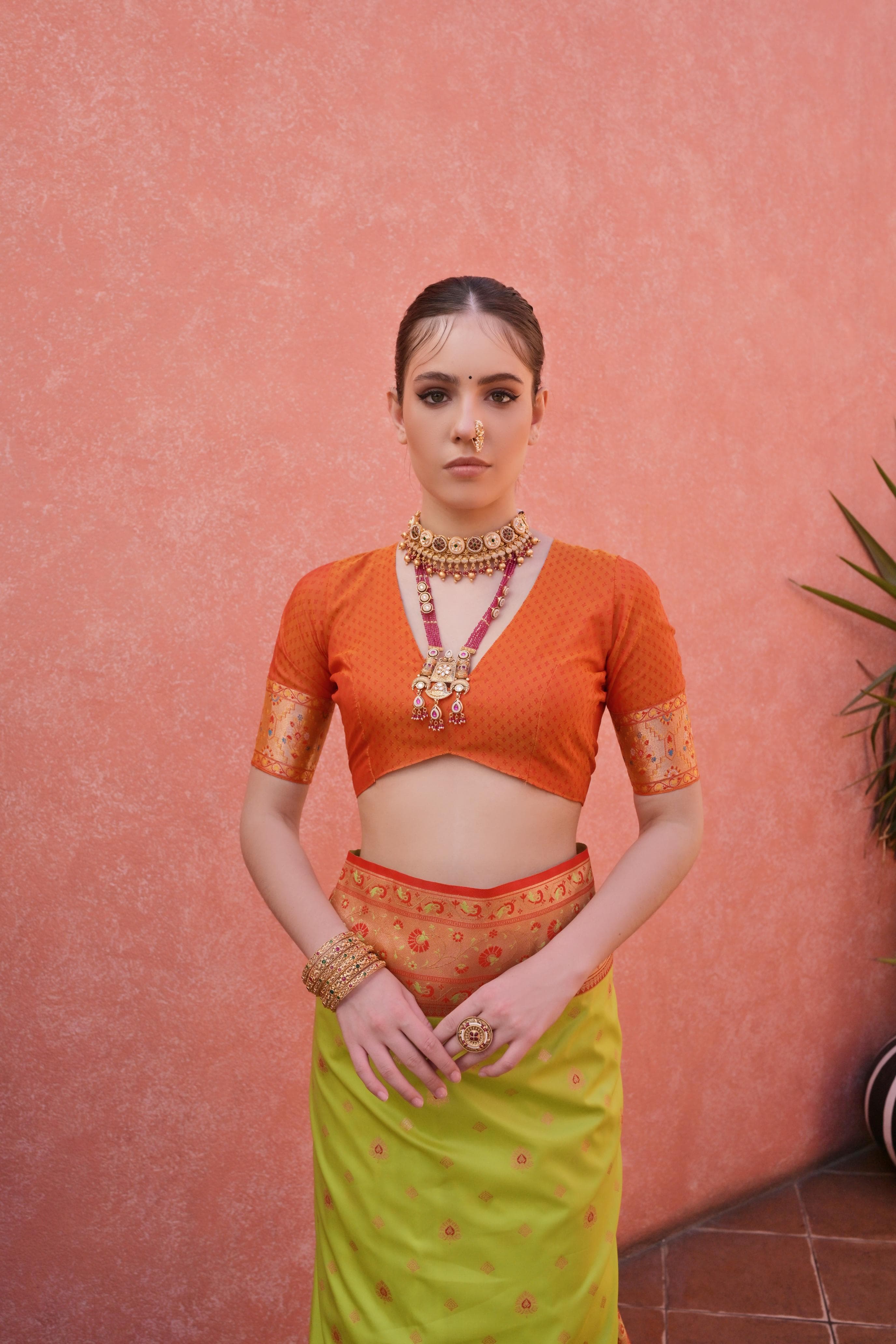 A woman models a vibrant Banarasi Silk Saree. The saree is lime green with fine Zari weaving, featuring a wide, ornate orange and red Zari woven border and pallu. She pairs it with a short-sleeve orange blouse and heavy traditional gold jewelry.