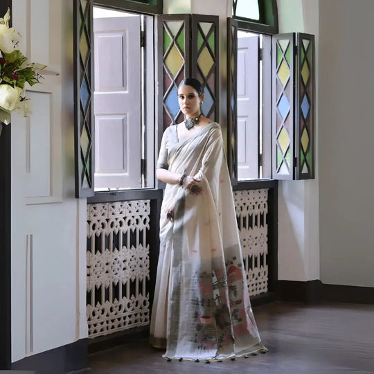 A woman models a graceful Off White Soft Muga Cotton Silk Saree. The saree has a thin grey border, and the pallu features a wide panel decorated with traditional Warli-style figurative motifs in black and grey. She wears a grey blouse and a dark oxidized choker necklace, posing indoors.