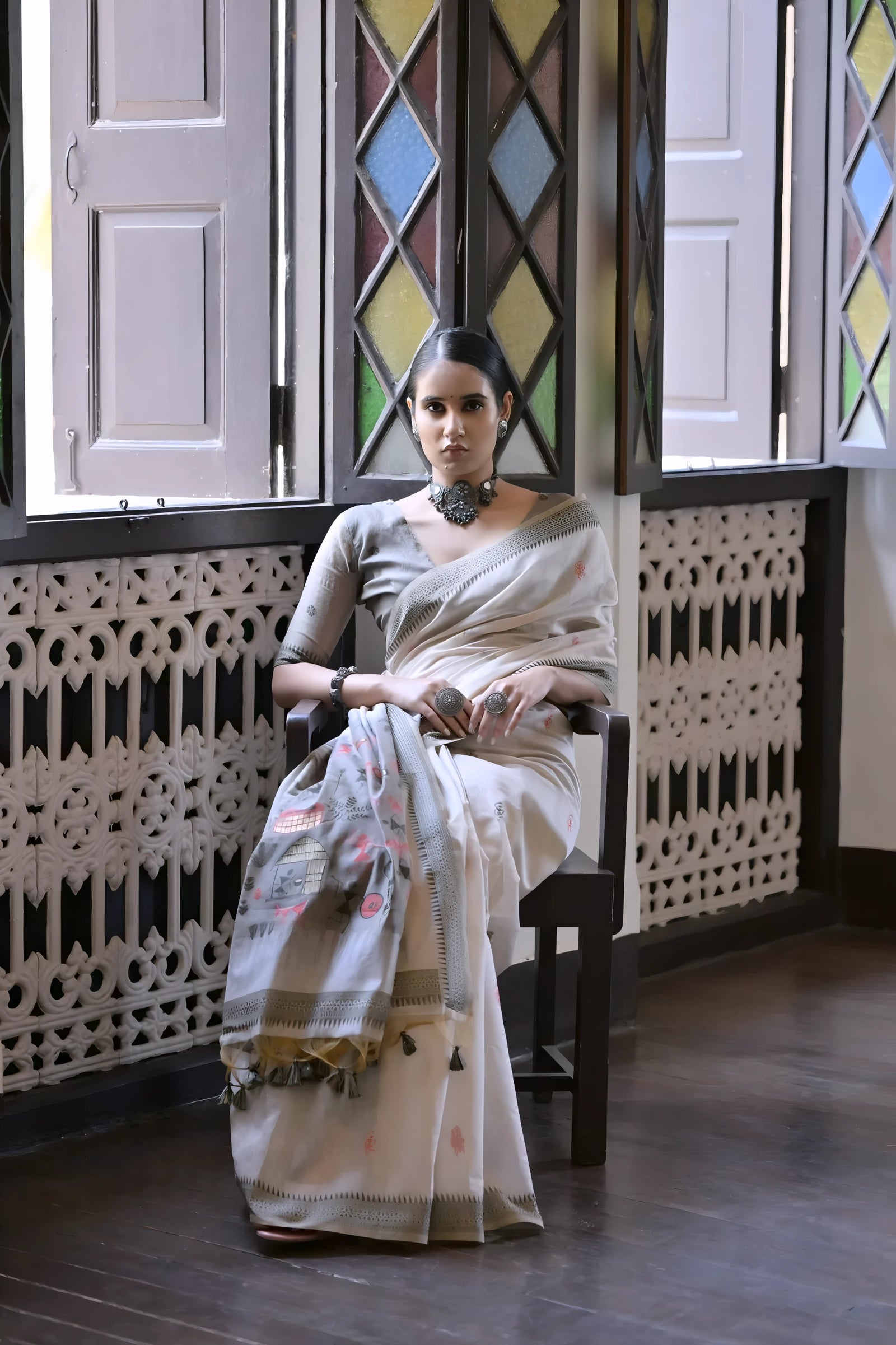 A woman models a graceful Off White Soft Muga Cotton Silk Saree. The saree has a thin grey border, and the pallu features a wide panel decorated with traditional Warli-style figurative motifs in black and grey. She wears a grey blouse and a dark oxidized choker necklace, posing indoors.