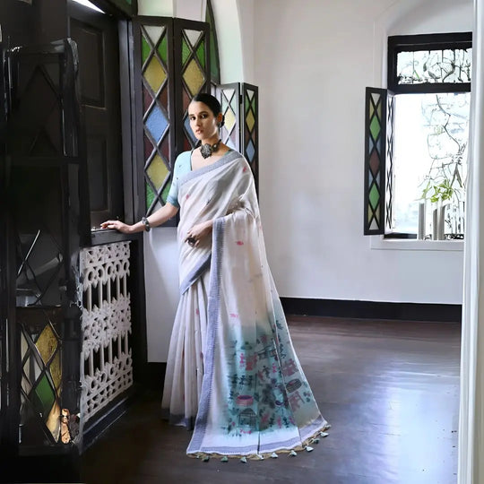 A woman models a graceful Off White Soft Muga Cotton Silk Saree. The pallu features a wide sea green panel decorated with traditional Warli-style figurative motifs in pink and red. She wears a light blue blouse and a dark oxidized choker necklace, posing indoors.