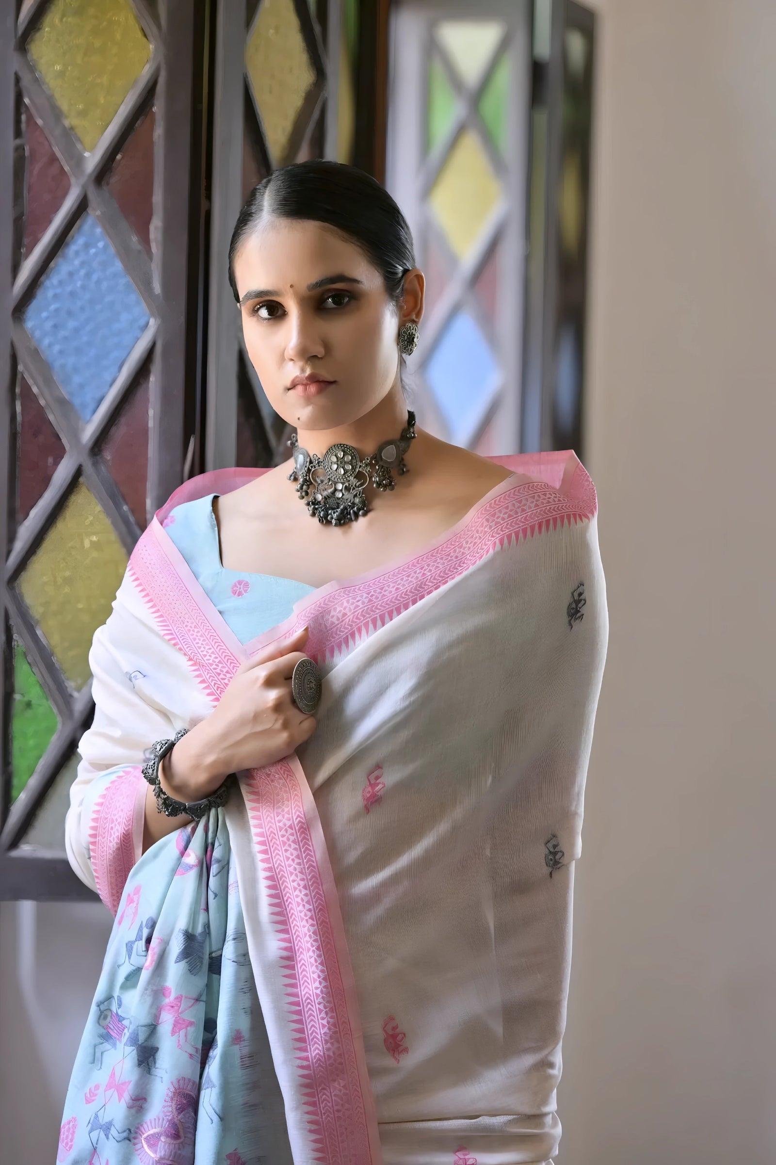 A woman models a graceful Off White Soft Muga Cotton Silk Saree. The saree has a dark pink woven border, and the pallu features a wide sky blue panel decorated with traditional Warli-style figurative motifs. She wears a light blue blouse and a dark oxidized  necklace, posing indoors.