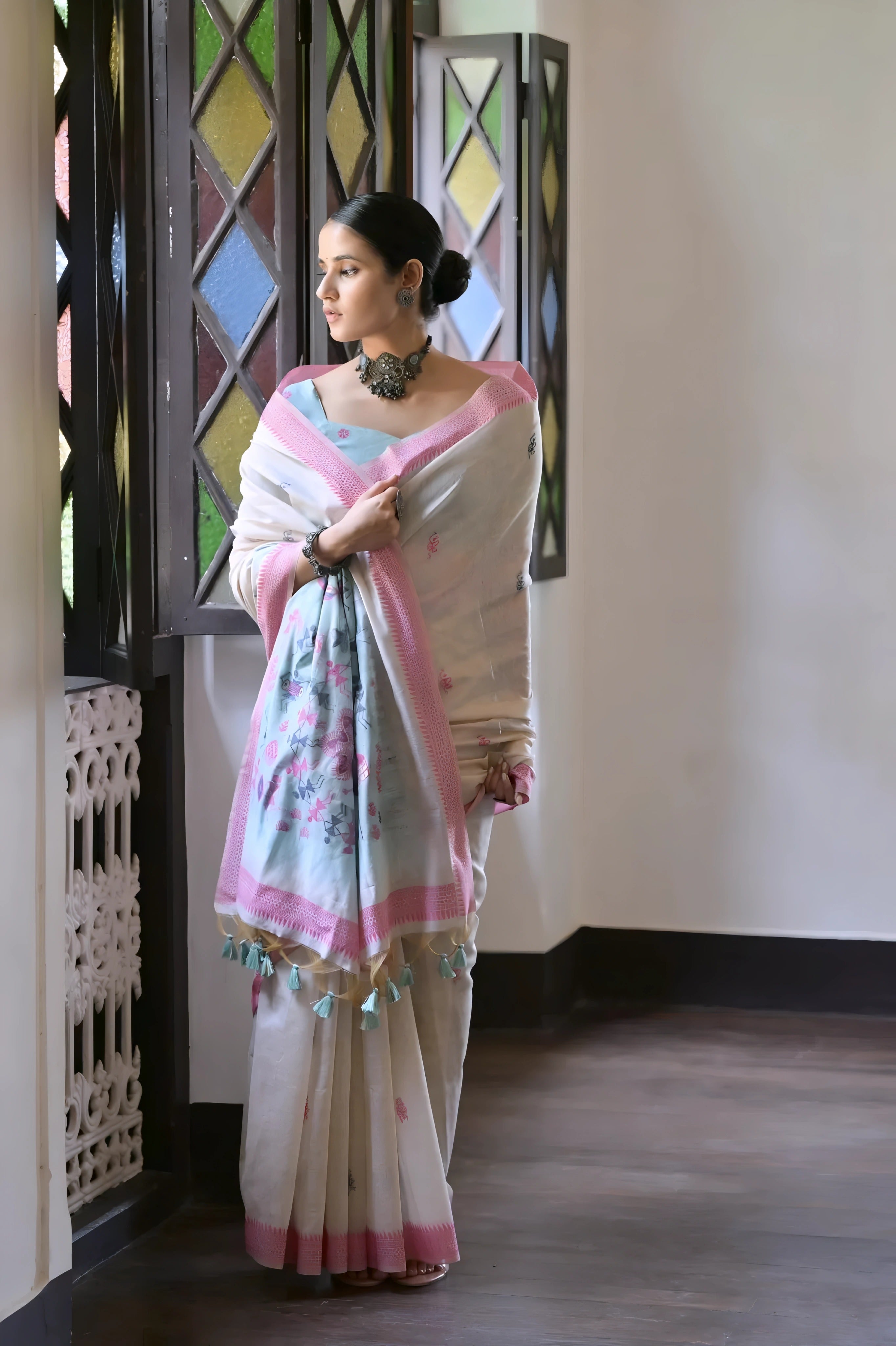 A woman models a graceful Off White Soft Muga Cotton Silk Saree. The saree has a dark pink woven border, and the pallu features a wide sky blue panel decorated with traditional Warli-style figurative motifs. She wears a light blue blouse and a dark oxidized choker necklace,  indoors.