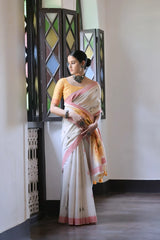 A woman models a graceful Off White Soft Muga Cotton Silk Saree. The saree has a pink accent border, and the pallu features a wide yellow panel decorated with traditional Warli-style figurative motifs. She wears a yellow blouse and a dark oxidized choker necklace, posing indoors.