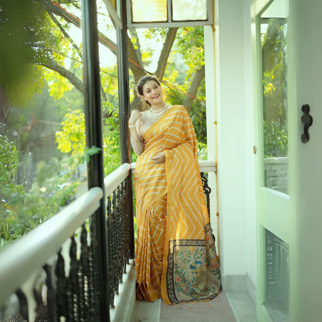 A woman models a vibrant Golden Yellow Saree with a white chevron/striped pattern. The pallu is beige with a dark border, featuring elaborate printed motifs of birds and flowers. She wears a pearl necklace and a lace-sleeve blouse, posing on a sunny balcony.