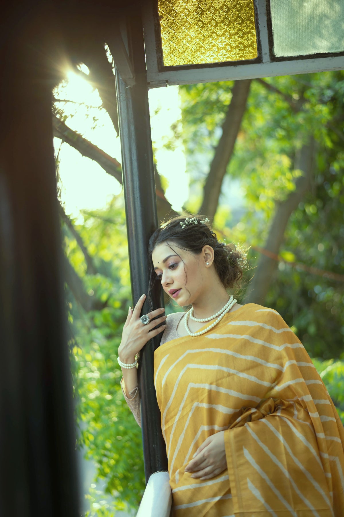 A woman models a vibrant Golden Yellow Saree with a white chevron/striped pattern. The pallu is beige with a dark border, featuring elaborate printed motifs of birds and flowers. She wears a pearl necklace and a lace-sleeve blouse, posing on a sunny balcony.