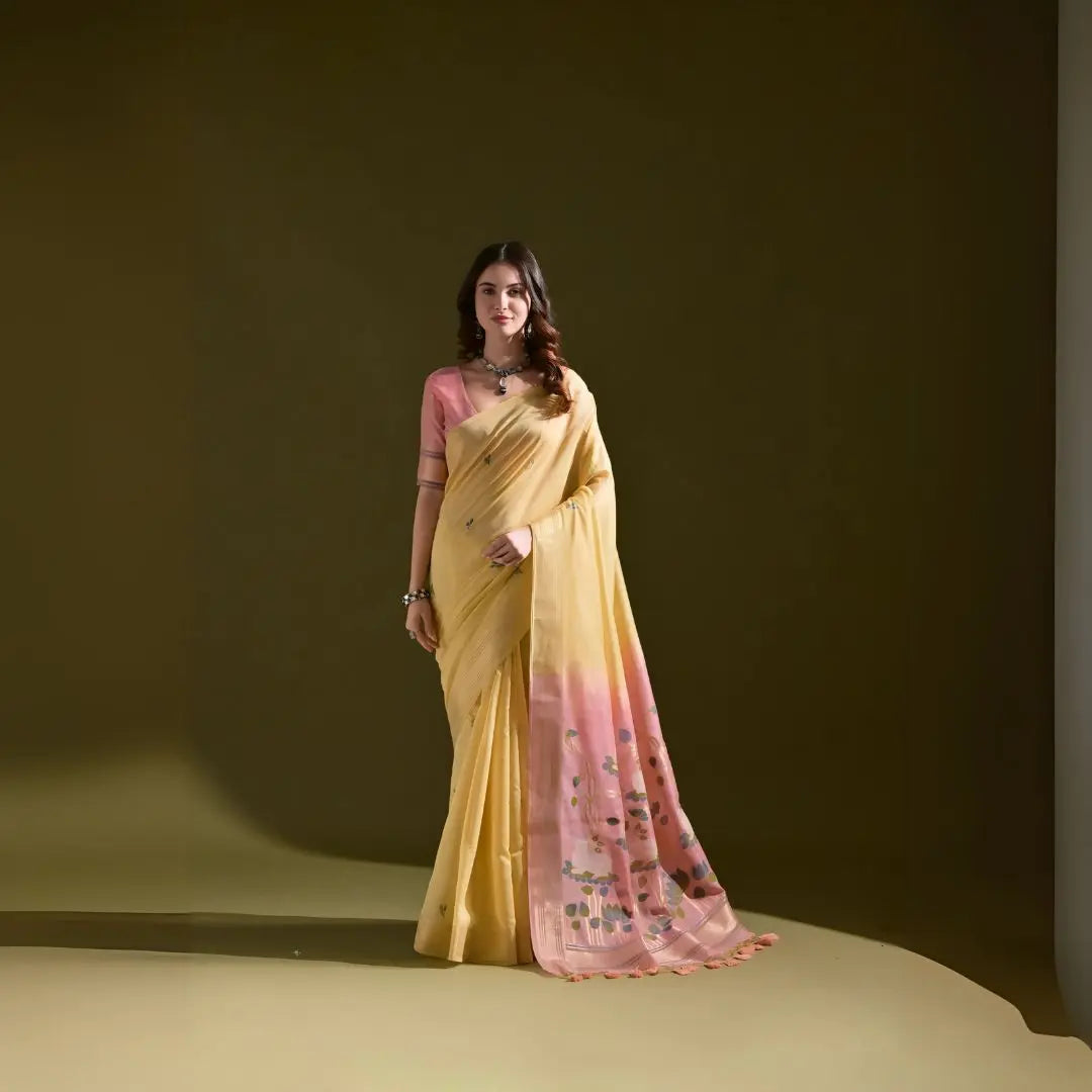  Woman wearing a yellow silk saree with pink floral print and matching pink blouse, standing against an olive green background.