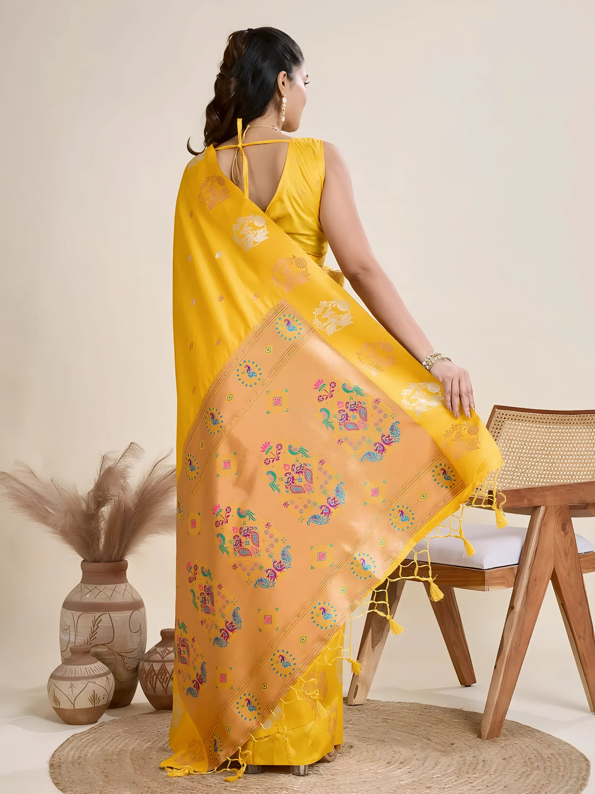 Woman wearing a vibrant yellow Banarasi silk saree with intricate pastel floral motifs and a contrasting gold border, accessorized with a statement necklace and bangles, elegantly posing beside a wooden chair in a minimalist studio setting with neutral decor, ceramic vases, and pampas grass.