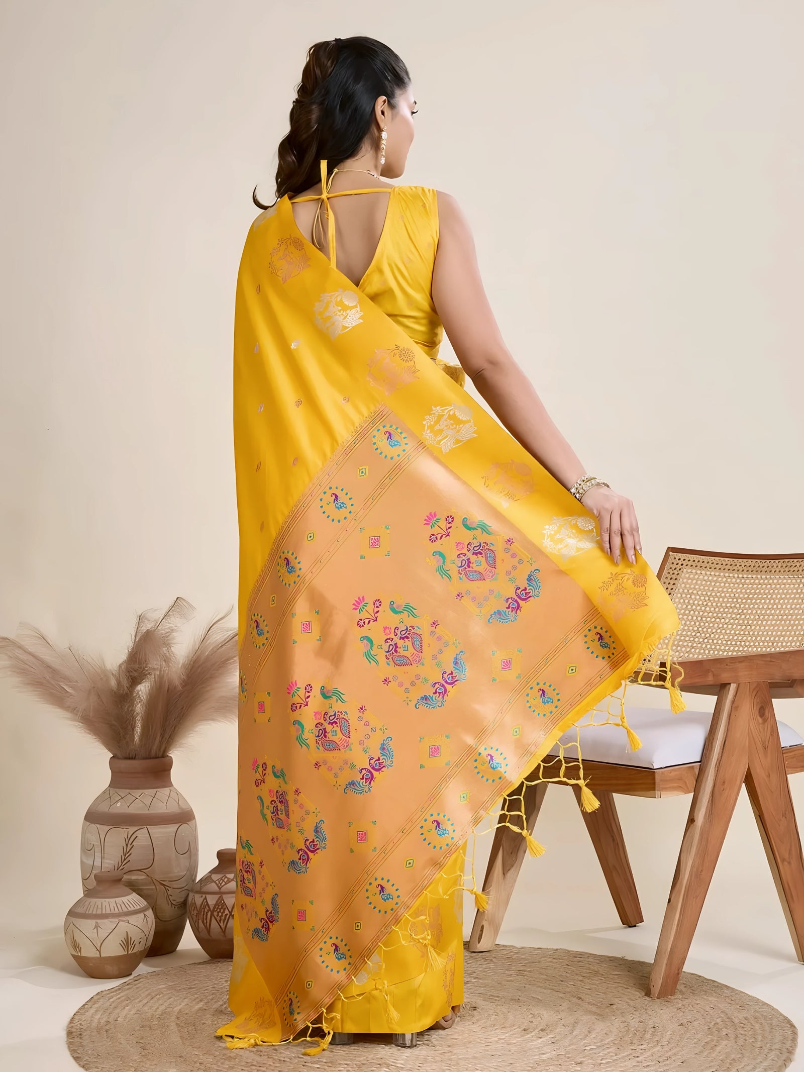 Woman wearing a vibrant yellow Banarasi silk saree with intricate pastel floral motifs and a contrasting gold border, accessorized with a statement necklace and bangles, elegantly posing beside a wooden chair in a minimalist studio setting with neutral decor, ceramic vases, and pampas grass.