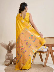 Woman wearing a vibrant yellow Banarasi silk saree with intricate pastel floral motifs and a contrasting gold border, accessorized with a statement necklace and bangles, elegantly posing beside a wooden chair in a minimalist studio setting with neutral decor, ceramic vases, and pampas grass.