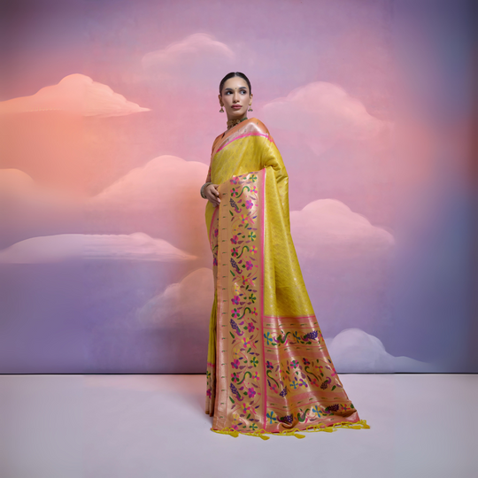 A woman models a luxurious Golden Yellow Soft Silk Saree with dense tone-on-tone weaving. The wide border and pallu feature heavy, multi-colored Paithani-style weaving in luminous peach, pink, purple, and green threads. She wears a soft pink blouse and a gold choker necklace, posing against a fantastical cloud backdrop.