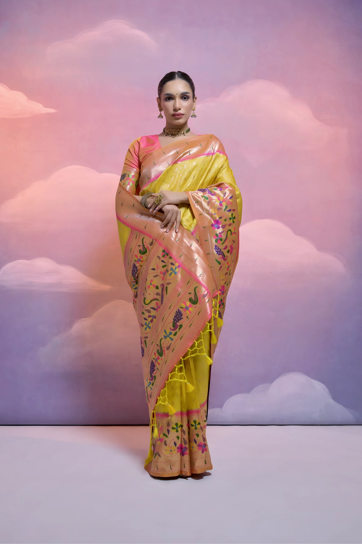 A woman models a luxurious Golden Yellow Soft Silk Saree with dense tone-on-tone weaving. The wide border and pallu feature heavy, multi-colored Paithani-style weaving in luminous peach, pink, purple, and green threads. She wears a soft pink blouse and a gold choker necklace, posing against a fantastical cloud backdrop.