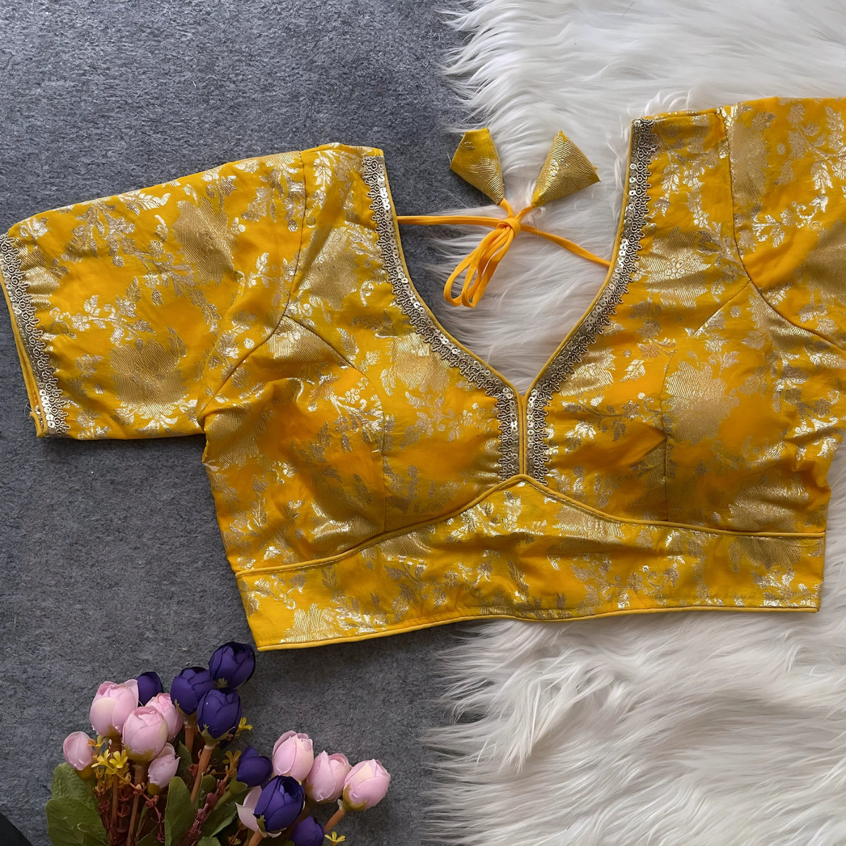  Yellow brocade blouse featuring intricate gold floral zari work, decorative trim along the neckline and sleeves, and a tie-up at the back, displayed flat on a grey surface with a white faux fur accent and a bunch of artificial pink and purple flowers in the corner, showcasing its festive ethnic design and elegant craftsmanship.