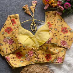 Yellow floral embroidered padded blouse with back tie detail, displayed on a grey surface with artificial flowers and white fur in the background.